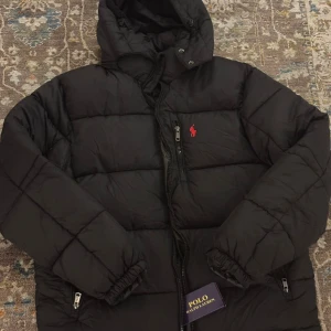 Svart pufferjacka från Polo Ralph Lauren - Svart pufferjacka från Polo Ralph Lauren med huva och dragkedja framtill. Jackan har ett rött Polo-logo broderat på bröstet och två sidofickor med dragkedja. Den är quiltad och har elastiska muddar vid ärmsluten för extra komfort.