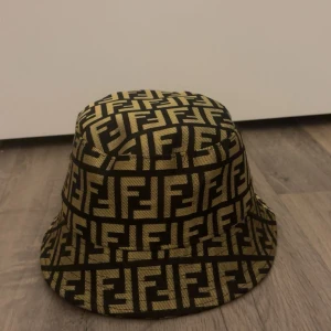 Fendi bucket hat med FF-mönster - Snygg bucket hat från Fendi med ikoniskt FF-logomönster i guld och svart. Hatten har en klassisk rund form och är tillverkad i ett slitstarkt tygmaterial. Perfekt för dig som vill sticka ut med en lyxig och trendig accessoar.