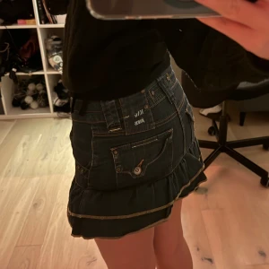 MNG Jeans volangkjol i denim - Supersöt kort jeanskjol från MNG Jeans med volangkant och snygga detaljer som fickor, bälteshällor och dragkedja fram. Kjolen är mörkblå med kontrastsömmar i gult och har en cool streetstil. Perfekt för dig som gillar denim och vill sticka ut.