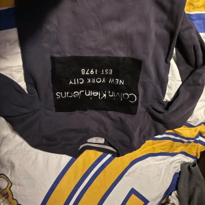 Mörkblå sweatshirt från Calvin Klein - Snygg mörkblå sweatshirt från Calvin Klein Jeans med stor svart patch och vit text på bröstet. Rund hals och långa ärmar, perfekt för dig som gillar streetstyle och vill ha en enkel men stilren look.