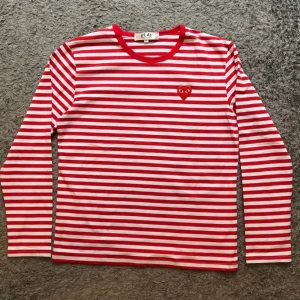 Striped Comme des Garçons longsleeve - Säljer en vit och röd randig långärmad tröja från Comme des Garçons PLAY. Tröjan har ett broderat rött hjärta med ögon på bröstet och rund halsringning. Tillverkad i mjuk bomull, perfekt för dig som gillar clean och lekfull stil.
