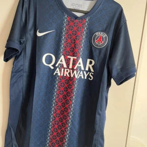 PSG matchtröja blå Nike - Säljer en officiell Paris Saint-Germain fotbollströja från Nike i marinblått med röd och vit grafisk rand på framsidan. Tröjan har korta ärmar, PSG-logga på bröstet och Qatar Airways som sponsor. Materialet är lätt och ventilerande polyester.