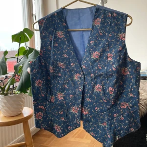 Blommig blå väst med knappar - Snygg väst i mörkblått tyg med blommigt mönster i rosa och grönt. Västen har klassisk V-ringning, tre blå knappar framtill och två fickor. Materialet känns som bomull och passformen är normal. Perfekt för dig som vill sticka ut med en cool vintage-vibe. 