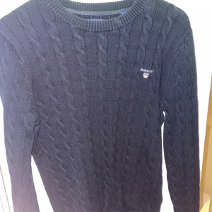 Mörkblå kabelstickad tröja från Gant - Kabelstickad mörkblå tröja från Gant med rund halsringning och broderad logga på bröstet. Tröjan har långa ärmar och ribbade muddar, perfekt för lager-på-lager under kyliga dagar.