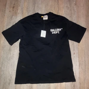  Gallery Dept t-shirt - Svart t-shirt från Gallery Dept. med vit tryckt logga på bröstet och stor text på ryggen med 'GALLERY DEPT. HOLLYWOOD, CA.'. Klassisk rund hals och korta ärmar. Materialet är mjuk bomull som känns skön mot huden. Perfekt för dig som gillar streetwear och tydliga prints. Storlek S men sitter som M/L