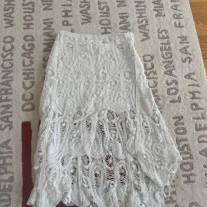 Vit långkjol med spets och volang - Superfin vit långkjol med detaljerad spets och volangkant. Kjolen har ett unikt virkat mönster med transparenta partier som ger en bohemisk vibe. Perfekt för dig som gillar romantiska och kreativa outfits. Materialet känns som bomull och spets, och kjolen har en flowy siluett.