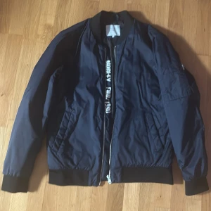 Mörkblå bomberjacka Jack & Jones L - Snygg mörkblå bomberjacka från Jack & Jones Core med svart ribbad krage, muddar och nederkant. Jackan har dragkedja framtill, fickor på sidorna och en ärmficka. Tryck med text på ryggen och insidan. Perfekt för dig som gillar streetstyle.