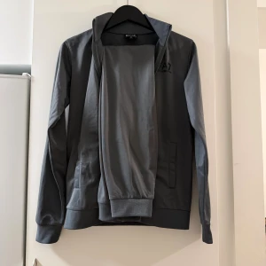 Grå/grön  zipjacka från EA7 Emporio Armani - Snygg grå zipjacka från EA7 Emporio Armani med diskret logga på bröstet. Jackan har hög krage, långa ärmar och ribbade muddar. Tillverkad i mjuk bomullsmix och stängs med dragkedja framtill. Perfekt för en clean och sportig look.