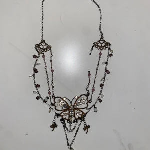 Vintage halsband med fjäril och pärlor - Unikt halsband med antikguld kedja, dekorerat med pärlor i vitt och rosa samt små glittriga stenar. Mittdelen har en stor fjäril med strass och hängande berlocker, bland annat små trollsländor. Romantisk och bohemisk vibe, perfekt för dig som gillar detaljer.  Ena kedjan på högra sidan var trasig (se bild 3) när jag köpte den second hand innan, men borde inte vara svårt att knipsa ihop 🤞🏽🤞🏽