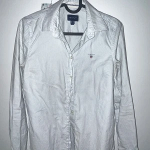 Vit randig skjorta från Gant - Snygg vit skjorta från Gant med tunna ljusblå ränder och klassisk krage. Skjortan har lång ärm, knappar framtill och en liten broderad logga på bröstet. Perfekt för dig som gillar stilrena och tidlösa plagg. Storlekar 34