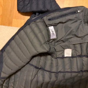 Mörkblå Colmar pufferjacka - Säljer en mörkblå pufferjacka från Colmar med quiltad design och dragkedja framtill. Jackan har långärmade ärmar, hög krage och två sidofickor med dragkedja. Materialet är lätt och syntetiskt, perfekt för kyliga dagar. Colmar-logga på kragen.