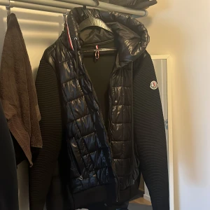 Svart pufferjacka från Moncler - Säljer en svart pufferjacka från Moncler med glansig quiltad framsida och huva med röd-vit-blå detalj. Ärmarna är ribbade och har Moncler-logga på vänster arm. Jackan har dragkedja och en modern, sportig look som passar perfekt till streetwear.