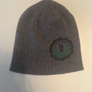 Grå ribbstickad mössa med brodyr från Stadium - Grå ribbstickad mössa från Stadium med svart och grön broderad logga framtill. Tillverkad i 100% akryl. I mycket bra skick.