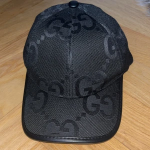 Svart keps från Gucci med GG-mönster - Snygg svart keps från Gucci med ikoniskt GG-mönster ton-i-ton över hela kepsen. Kepsen har böjd skärm med läderdetalj längs kanten och justerbart spänne bak i svart läder. Tillverkad i bomull och polyester för en exklusiv känsla.