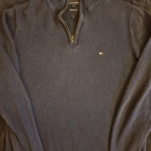 Mörkblå half zip tröja Tommy Hilfiger - Snygg mörkblå långärmad tröja från Tommy Hilfiger med half zip och liten broderad logga på bröstet. Tröjan är gjord i premium bomull och har ribbade muddar vid hals, ärmslut och nederkant. Perfekt för dig som gillar stilrena och klassiska plagg.