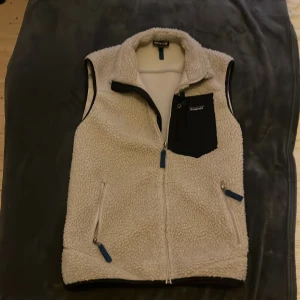 Beige fleeceväst från Patagonia - Mysig beige fleeceväst från Patagonia med hög krage, två sidofickor med dragkedja och en svart bröstficka. Västen har blå detaljer på dragkedjorna och svart kantband. Perfekt för lager-på-lager och outdoor-vibe.