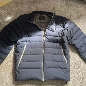 Mörkblå dunjacka från Sail Racing - Säljer en mörkblå dunjacka från Sail Racing i storlek medium. Jackan har quiltad design, ljusgrått foder och dragkedjor i kontrastfärg. Perfekt för kyliga dagar och har en clean look med Sail Racing-logga på bröstet.