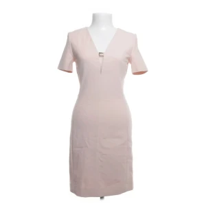 Ljusrosa miniklänning med v-ringning och strass - En stilren ljusrosa kortklänning med v-ringning och dekorativ metall-detalj framtill. Klänningen har korta ärmar och en figurnära passform som ger en elegant siluett. Perfekt för dig som gillar minimalistisk och chic stil.