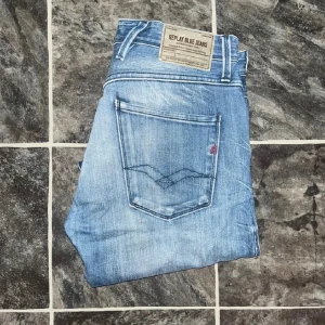 Replay Jeans - Snygga ljusblå Replay Anbass jeans med klassisk femficksdesign och diskreta slitningar framtill. Jeansen har röda sömmar på insidan och en slim passform som sitter tajt längs benen. Midja 38cm, längd 99cm. JAG SKICKAR EJ FLER BILDER NÄR JAG BÄR JEANSEN! för storleksguide hänvisar jag till måtten och för bättre inblick i passformen rekommenderar jag att googla på modellnamnet. Jeansen kan vara uppsydda utan att jag noterat det så kolla måtten noggrant :) s4