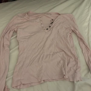 Ljusrosa ribbad långärmad tröja Lindex - Säljer en ljusrosa långärmad tröja från Lindex med ribbad struktur och v-ringning med dekorativa knappar. Tröjan är mjuk och har en slim passform som sitter snyggt. Perfekt för dig som gillar enkel och stilren look.