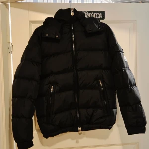 Svart Moncler pufferjacka med huva - Svart pufferjacka från Moncler med huva och två sidofickor med dragkedja. Jackan har quiltad design, lång ärm och coolt Moncler-märke på ärmen. Insidan har ett serietidningsmotiv och flera etiketter. Perfekt för kalla dagar. Priset går att diskuteras vid en snabb affär.