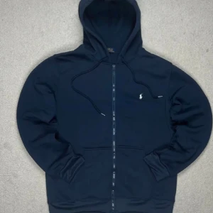 Marinblå hoodie från Polo Ralph Lauren - Snygg marinblå hoodie från Polo Ralph Lauren med dragkedja och huva med snörning. Klassisk vit broderad logga på bröstet. Tröjan har långa ärmar och praktiska fickor framtill. Perfekt för chill dagar och streetwear-stil.