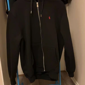 Svart hoodie från Polo Ralph Lauren - Svart hoodie från Polo Ralph Lauren med röd logobrodyr på bröstet. Tröjan har dragkedja framtill, snörning i huvan och en stor magficka. Tillverkad i mjuk bomull och har långa ärmar med ribbade muddar. Perfekt för chill dagar.