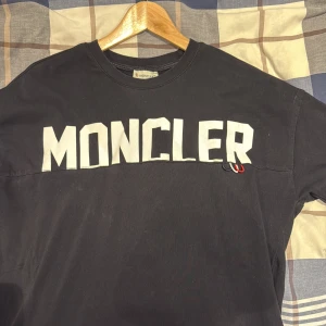 Svart Moncler t-shirt med stor logga - Svart t-shirt från Moncler med stor vit logga över bröstet och en massiv vit M på ryggen. Klassisk rund hals och korta ärmar. Materialet är mjuk bomull som känns skönt mot huden. Perfekt för dig som gillar streetwear och tydliga prints.