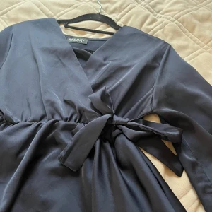 Svart omlottblus med knyt från Missay - Snygg svart omlottblus från Missay med långa ärmar och v-ringad hals. Blusen har ett brett knytband i midjan som ger en cool siluett och extra detalj. Materialet är mjukt och har en lätt glansig finish som känns lyxig. Perfekt för dig som gillar stilrena plagg med twist.