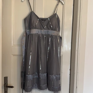 Grå paljettklänning från ONLY - Supersnygg grå kortklänning från ONLY med tunna axelband och v-ringad omlott framtill. Klänningen har glittriga paljettränder och ett brett satinband i midjan. Materialet är polyester och satin, perfekt för att sticka ut med lite extra bling.