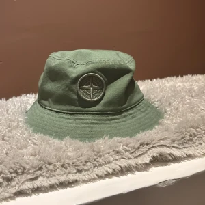 Grön bucket hat från Stone Island - Snygg grön bucket hat från Stone Island med klassisk broderad kompasslogga framtill. Hatten är tillverkad i slitstark bomull och har en rundad form med bred brätte. Köpte på NK i Göteborg. Ny pris 1000kr