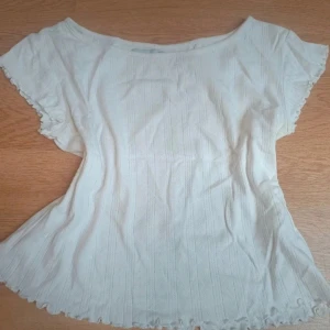 Vit blus med volangkant ifrån lager 157 - Säljer en söt vit blus med korta ärmar och volangkant vid ärmslut och nederkant. Blusen har ett diskret randigt mönster och är lätt figursydd för en feminin siluett. Perfekt för dig som gillar ljusa och fräscha toppar.