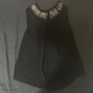 Svart blus med silvriga detaljer - Svart blus utan ärmar med rund halsringning dekorerad med silvriga stavar och silvriga knappar längs ryggen