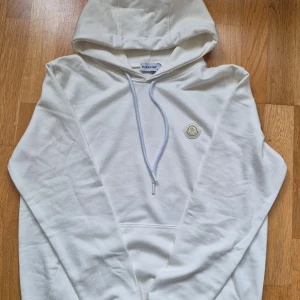 Vit hoodie från Moncler med logga - Snygg vit hoodie från Moncler i mjuk bomull. Tröjan har huva med snörning, klassisk Moncler-logga på bröstet och en stor magficka. Perfekt för dig som gillar stilrena och exklusiva plagg med streetkänsla.