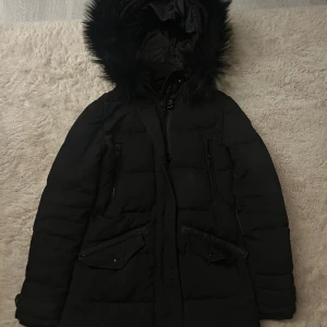 Svart Rockandblue dunjacka med päls - Svart lång dunjacka från Rockandblue med fluffig huva och snygga detaljer. Jackan har dragkedja, knappar, två stora fickor med lock och två snedställda fickor med dragkedja. Insidan är fodrad och huvan har pälsimitation. Perfekt för kalla dagar.