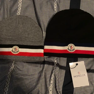 Moncler mössa med randig kant - Två snygga Moncler mössor, en i grått och en i svart. Båda har breda ränder i rött, vitt och svart eller blått nertill samt Moncler-logga framtill. Klassisk, tight passform och mjukt material som känns lyxigt. Perfekt accessoar för att lyfta din vinterstil. En Mössa för 400kr 