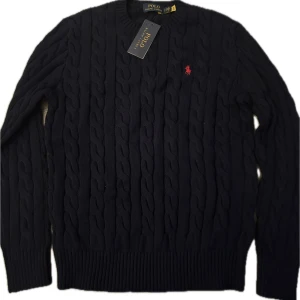 Mörkblå stickad tröja Polo Ralph Lauren - Snygg mörkblå stickad tröja från Polo Ralph Lauren med klassiskt kabelstickat mönster och röd broderad logga på bröstet. Tröjan har rund hals och långa ärmar, perfekt för dig som gillar stilren och tidlös design.