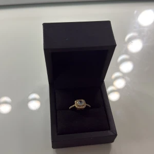 Guldig ring med glittrande stenar - Superfin ring i guldton med fyrkantig infattning och en stor glittrande sten i mitten, omgiven av mindre stenar. Ringen har även små stenar längs bandet för extra bling. Perfekt för dig som gillar att sticka ut med elegans.