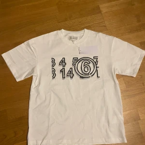 MM6 vit t-shirt - ! Pris går att diskuteras vid snabb affär ! MM6 t-shirt i vit bomull med svart grafiskt tryck av siffror på bröstet. Klassisk rund hals och korta ärmar. Snygg och clean design med märkets ikoniska etikett i nacken. Perfekt för dig som gillar streetwear och designerplagg.