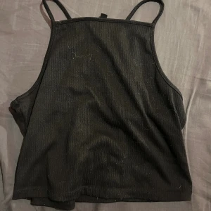 Svart ribbad linnetopp - Säljer en svart ribbad linnetopp med smala axelband och hög halsringning. Toppen är croppad och har en stilren, enkel design som funkar till många olika looks. Materialet är mjukt och stretchigt, perfekt för dig som gillar minimalistisk stil.