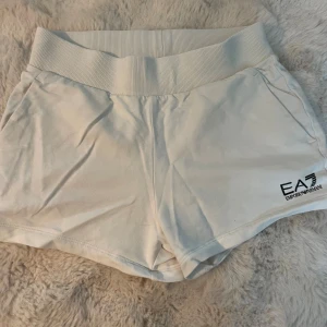 Vita shorts från Emporio Armani EA7 - Snygga vita shorts från Emporio Armani EA7 med bred ribbad midja och diskret logga framtill. Mjukt bomullsmaterial och fickor i sidorna gör dem både bekväma och stilrena. Perfekta för en chill och sportig look.