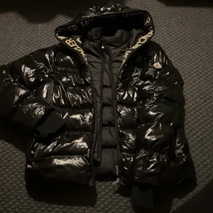 Svart Moncler pufferjacka med huva - Säljer en svart pufferjacka från Moncler i storlek L. Jackan har glansig finish, huva med Moncler-logga längs kanten och dragkedja framtill. Klassisk Moncler-patch på ärmen och breda quiltade partier. Perfekt för kalla dagar.