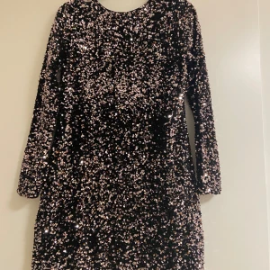 Svart paljettklänning med lång ärm - Glittrig svart kortklänning täckt av paljetter med lång ärm och rund halsringning. Klänningen har en snygg passform och en rosett i nacken som detalj. Perfekt för dig som vill sticka ut med en riktigt festlig look. Storlek 170 men passar som Xxs-xs. Använd 2 gånger