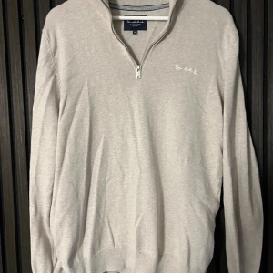  stickad tröja från Bondelid - Snygg beige stickad tröja från Bondelid med halv dragkedja och liten broderad logga på bröstet. Tröjan har långärmad passform och ribbade muddar vid ärmslut och nederkant. Perfekt för dig som gillar stilrena och klassiska plagg.