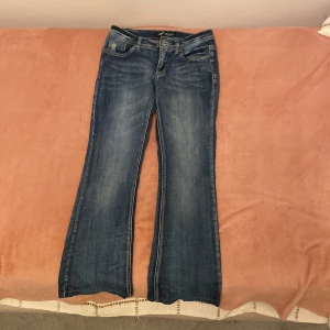 Blå bootcut jeans från Arizona - Snygga blå jeans från Arizona med bootcut passform och klassisk femficksdesign. Jeansen har slitningar och kontrastsömmar, samt detaljerade bakfickor med bruna broderier. Materialet är robust denim och midjan är normal.
