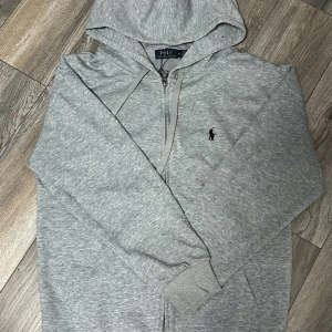 Grå hoodie från Polo Ralph Lauren - Snygg grå hoodie från Polo Ralph Lauren med dragkedja och huva. Tröjan har det klassiska Polo-logot broderat på bröstet och ribbade muddar vid ärmslut. Tillverkad i mjuk bomull, perfekt för chill dagar. Storlek M.