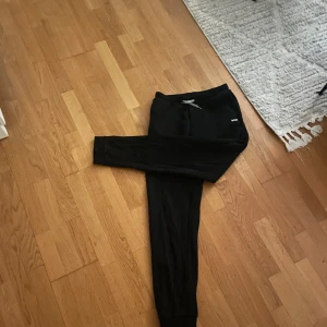 Svarta mjukisbyxor från BOSS - Svarta mjukisbyxor från BOSS med vit logga på vänster ficka. Byxorna har en slim passform med muddar vid bensluten och elastisk midja. Perfekta för chill eller träning, tillverkade i mjukt bomullsmaterial för extra komfort. Sitter som stl S