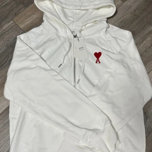 Vit hoodie med rött hjärta från Ami - Vit hoodie från Ami med dragkedja och huva. På bröstet finns ett broderat rött hjärta med bokstaven A. Tröjan har snörning vid huvan och är långärmad. Perfekt för dig som gillar stilrena detaljer och vill ha en chill look.