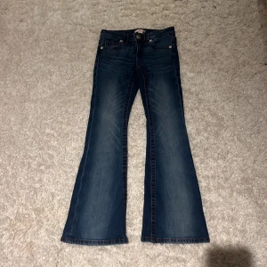 Blå bootcut jeans från Gina Tricot - Snygga blå jeans från Gina Tricot i bootcut-modell med röda kontrastsömmar och broderade detaljer på bakfickorna. Jeansen har klassisk femficksdesign och knappstängning framtill. Perfekt för dig som gillar retrovibbar och coola detaljer.