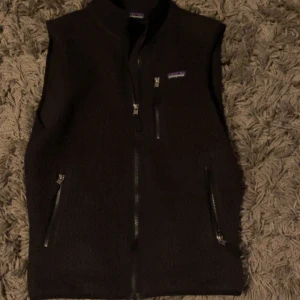 Svart fleeceväst från Patagonia - Svart fleeceväst från Patagonia i storlek S. Västen har hög krage, tre praktiska fickor med dragkedja och en stilren design utan mönster. Perfekt för lager-på-lager och outdoor-looks. Mjuk och varm fleece som håller dig cozy under kyliga dagar. Nypris 1300 använd max 5 gånger